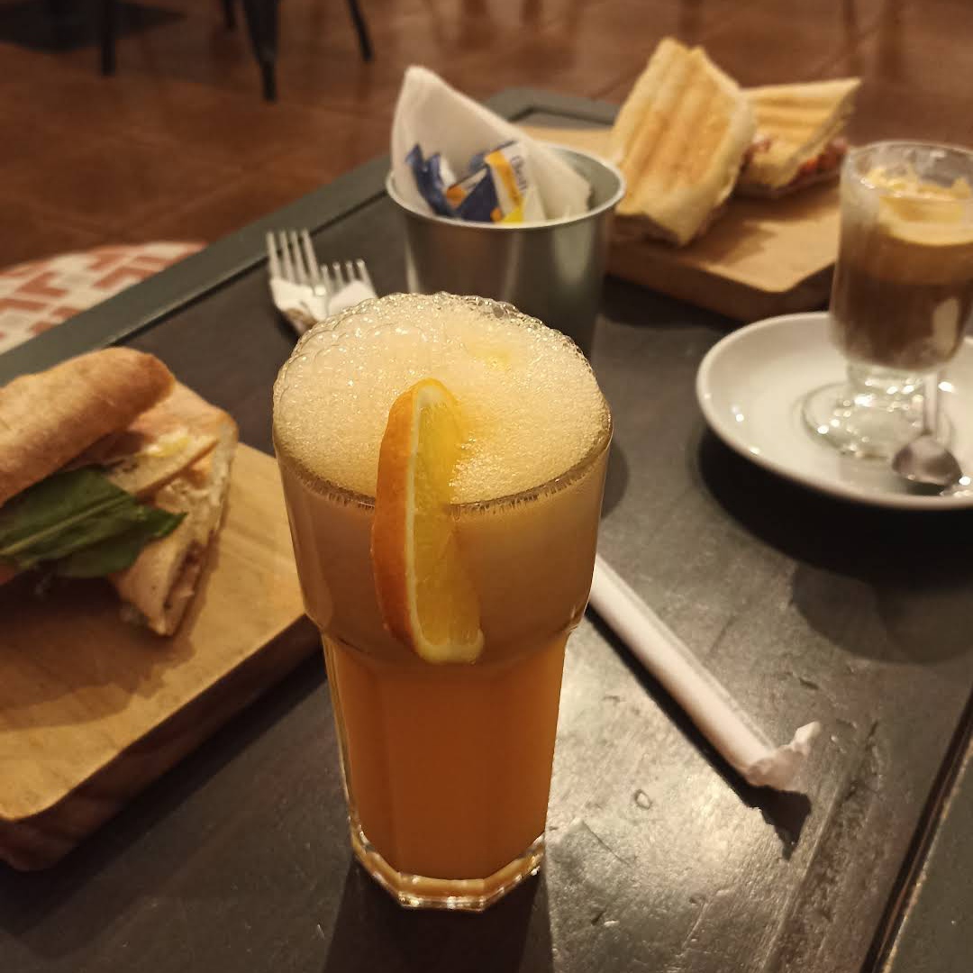 Imagen donde se muestra un jugo de naranja en primer plano y sanguches de fondo, cafetería Anticafé